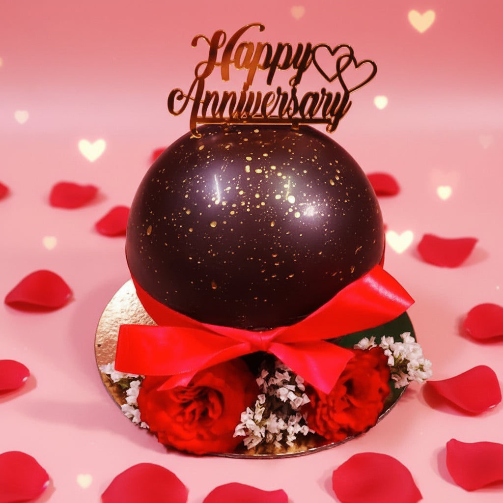 Decorative chocolate sphere with red ribbon and flowers, topped with 'Happy Anniversary' text on a wooden surface.