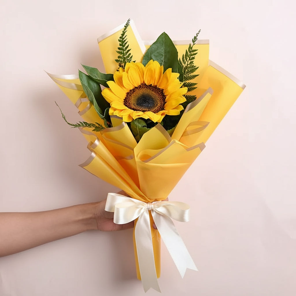 Bouquet of sunflowers wrapped in yellow paper with a black ribbon held by a hand on a light background